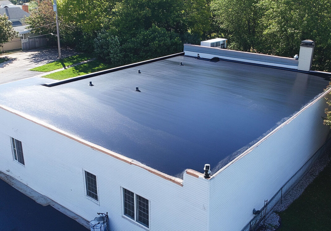 Freshly coated flat roof on commercial building in sunny weather Flat black roof on white building with trees nearby.